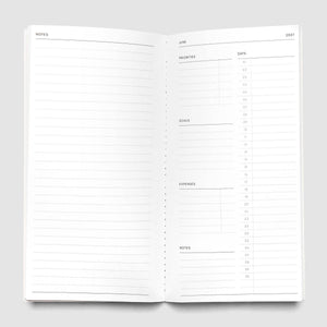 An open spiral-bound planner or notebook showing two white pages with minimalist black printed schedules and tables, photographed on a light gray background.