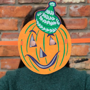 Person wearing a Halloween-themed pumpkin mask with a green hat against a brick wall.