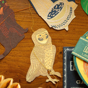 A collection of handcrafted origami paper models and books arranged on a wooden table. The central subject is a detailed owl-shaped origami model, surrounded by other paper creations including a lion, a bird, and a pinecone, with a book titled 'EAGLE SCREENPRINTS WITH SIDES' visible on the right.