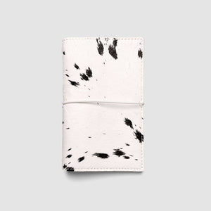 A folded white leather card holder or wallet with a black cow print pattern, shown on a neutral light gray background.