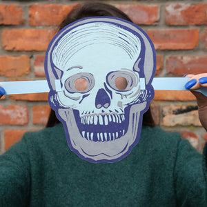 Person holding a skull-shaped mask over their face against a brick wall.