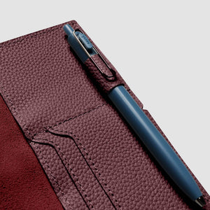 A close-up of a dark burgundy pebbled leather notebook cover with a blue pen tucked into a side pocket, set against a light gray background.