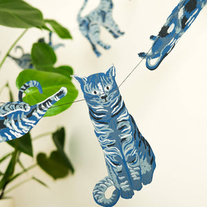 Blue and white cat-shaped hanging decorations strung across a room, with a potted plant and textured chair visible in the background.