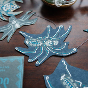 A close-up of a handmade paper garland featuring stylized blue and white paper bats, arranged on a dark wooden tabletop with a small bowl of wrapped stones in the background.