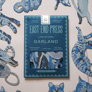 East End Press Garland hand-painted paper cutters displayed on a light beige wall, with the blue patterned cutters arranged around a central blue book cover.