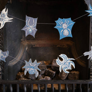 Spider-themed paper garland hanging in front of a dark fireplace. The garland features light blue and white spider silhouettes with web details.