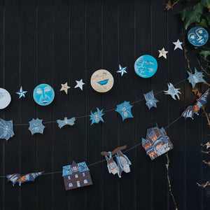 A decorative string garland with moon, star, and small haunted house cutouts in blue, beige, and brown tones, hung against a dark paneled wall with ivy on the right side.